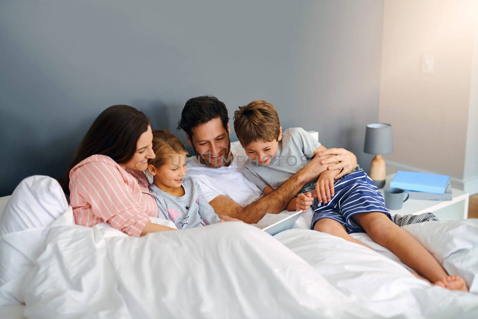 Waking up in the morning and spending time with family. a young family using a tablet while chilling in bed together at home. by YuriArcurs