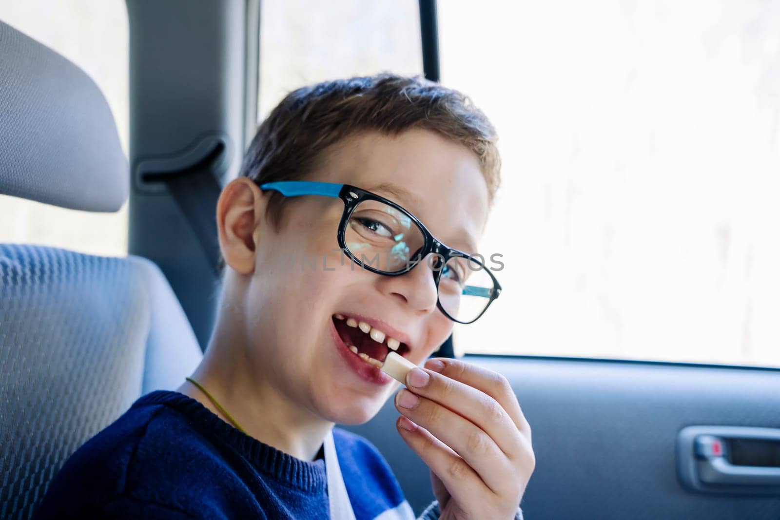 A happy Caucasian boy of school age rides in the back seat of a car and eats candy. A schoolboy with glasses. The child is wearing seat belts, and he is traveling with his family by car