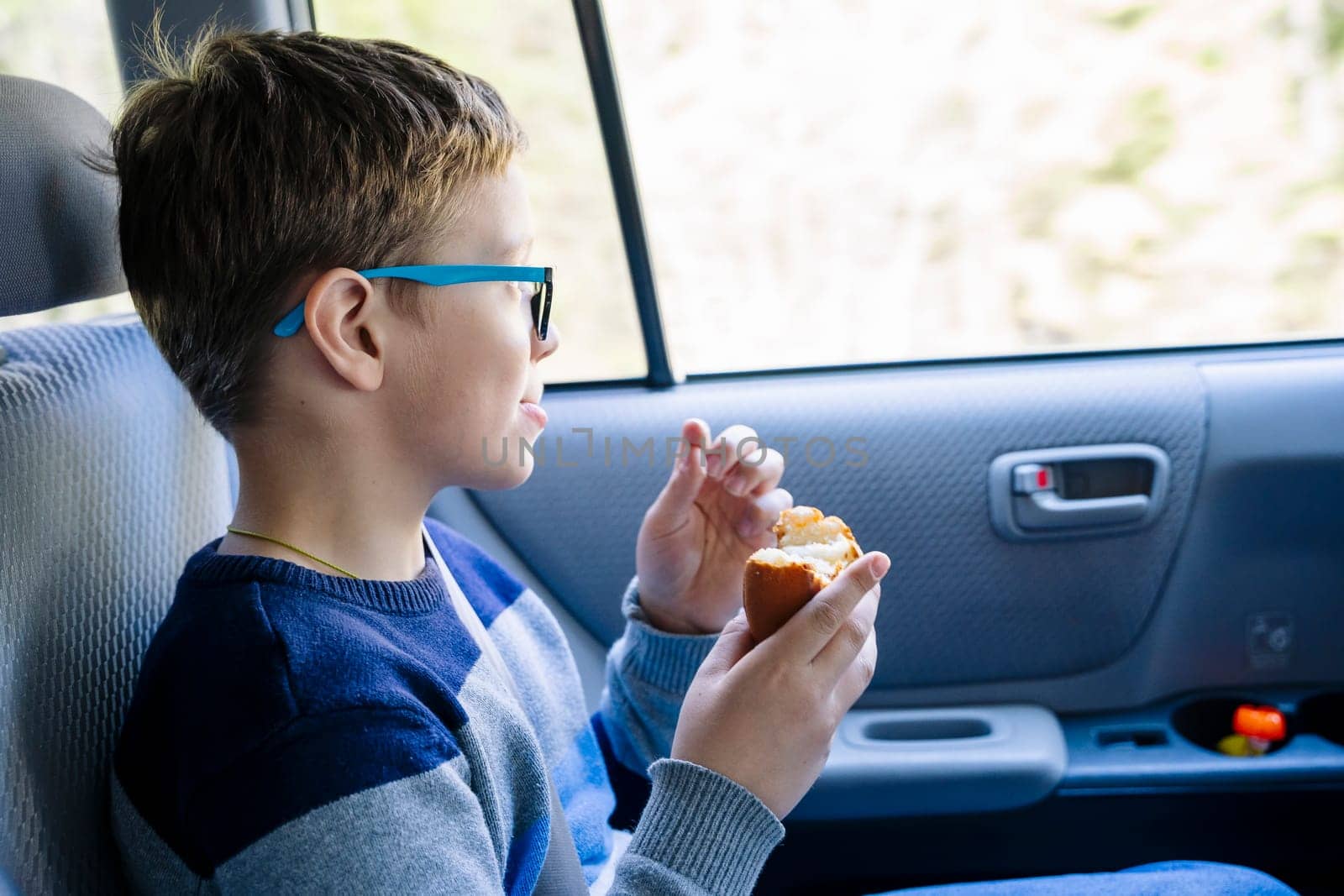 A happy Caucasian boy of school age rides in the back seat of a car and eats a sandwich. A schoolboy with glasses. The child is wearing seat belts, and he is traveling with his family by car