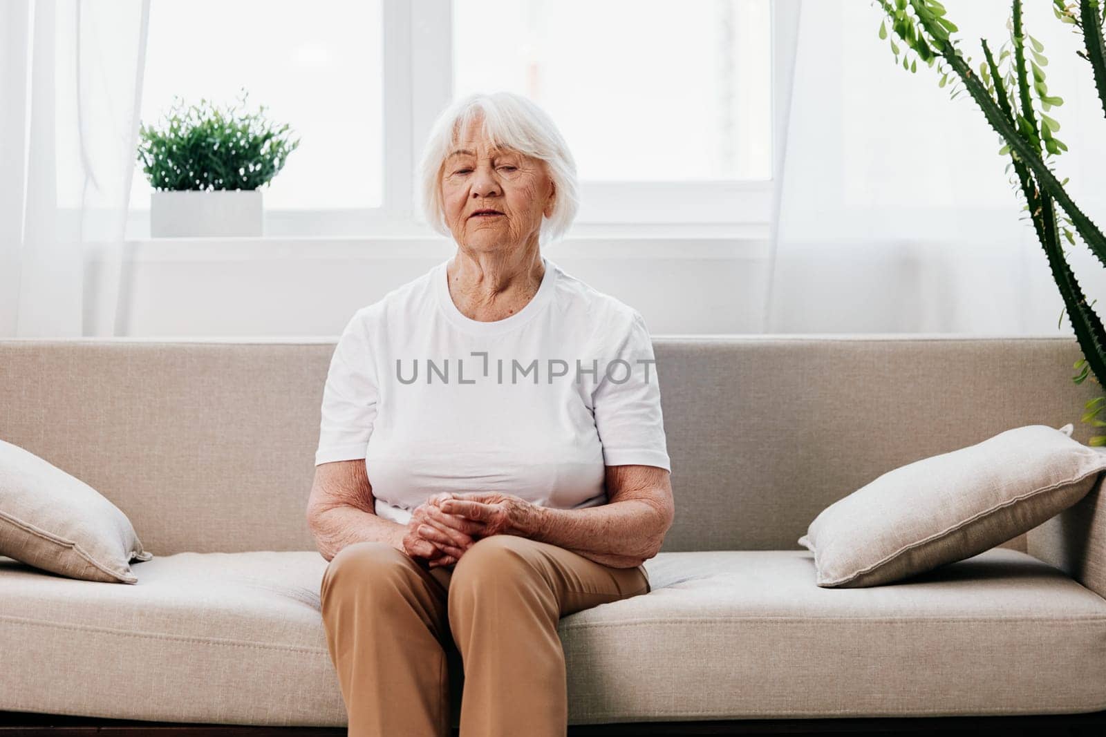 Elderly woman sits on sofa at home, bright spacious interior in old age smile, lifestyle. Grandmother with gray hair in a white T-shirt and beige trousers. by SHOTPRIME
