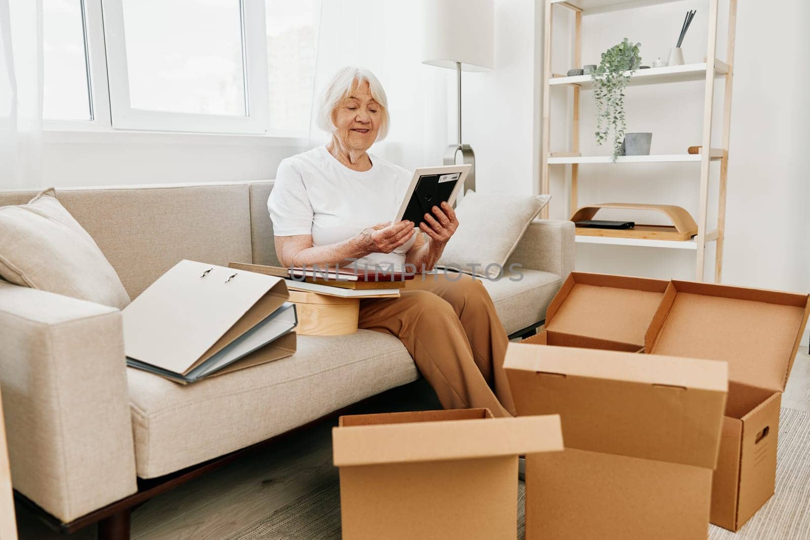 elderly woman sits on a sofa at home with boxes. collecting things with memories albums with photos and photo frames moving to a new place cleaning things and a happy smile. High quality photo