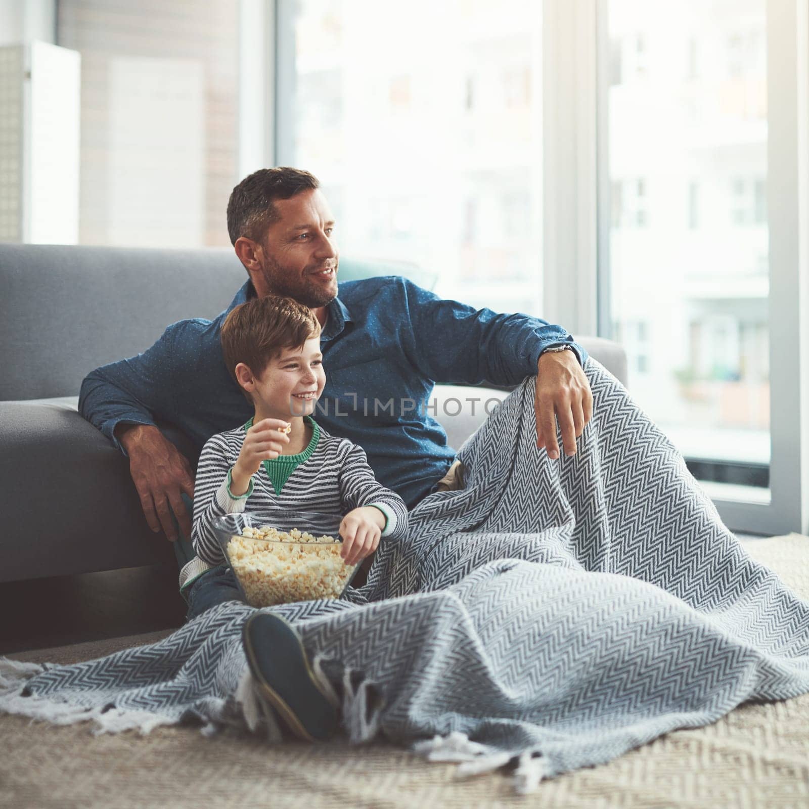 Thanks for the popcorn dad. a carefree young boy and his father watching a movie together while being seated on the floor at home during the day