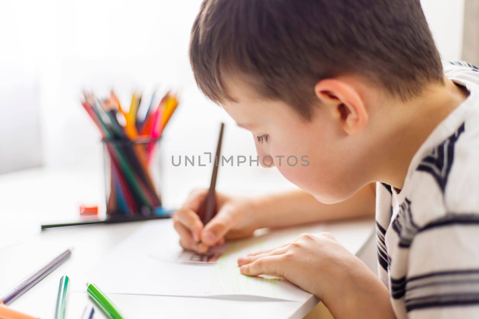 A child boy draws on white paper with colored pencils while sitting at a table.