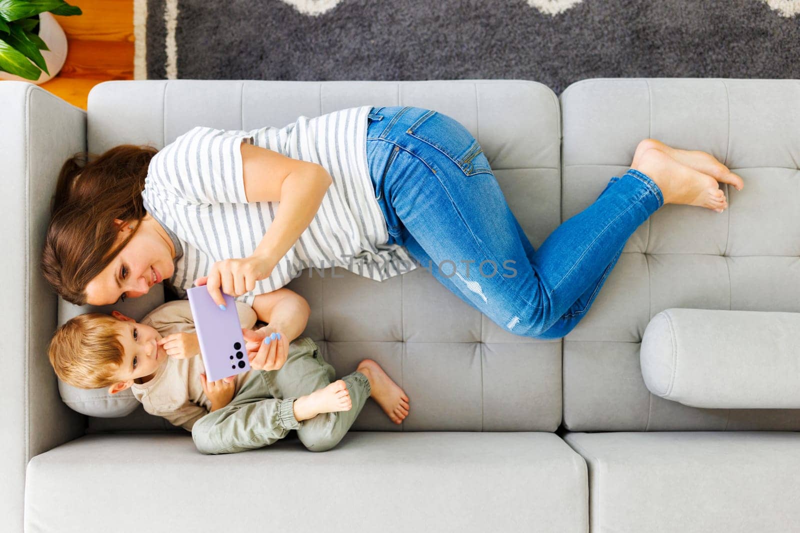 Woman and her little son lying on cozy sofa with smartphone, above view. Family spend leisure together at home using smartphone, play games, watch video, take selfies