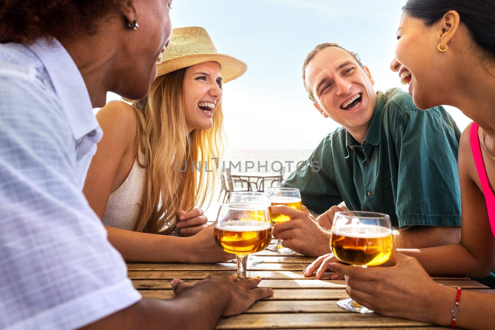 Group of multiracial young friends laughing having fun at beach bar drinking beers together. Friends on summer vacation. by Hoverstock