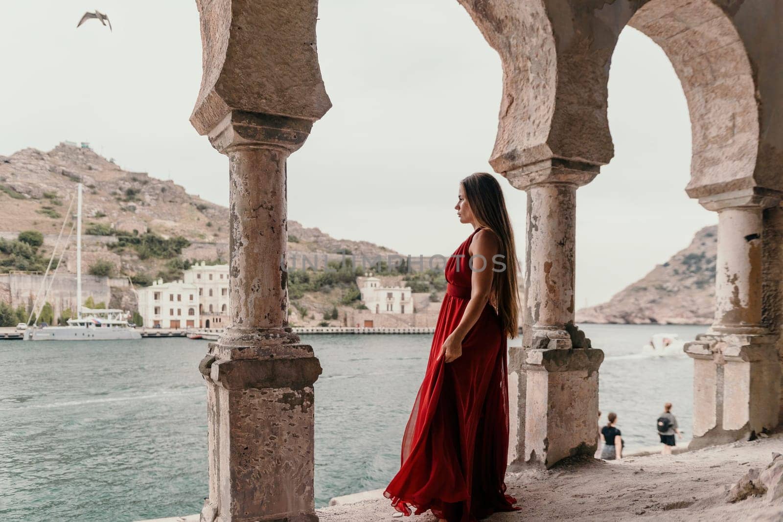 Side view a Young beautiful sensual woman in a red long dress posing on a volcanic rock high above the sea during sunset. Girl on the nature on overcast sky background. Fashion photo