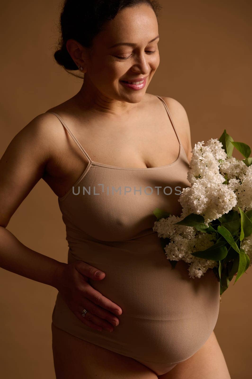 Fashion portrait pregnant woman in lingerie, holding white lilacs, stroking her belly, smiling over isolated background by artgf