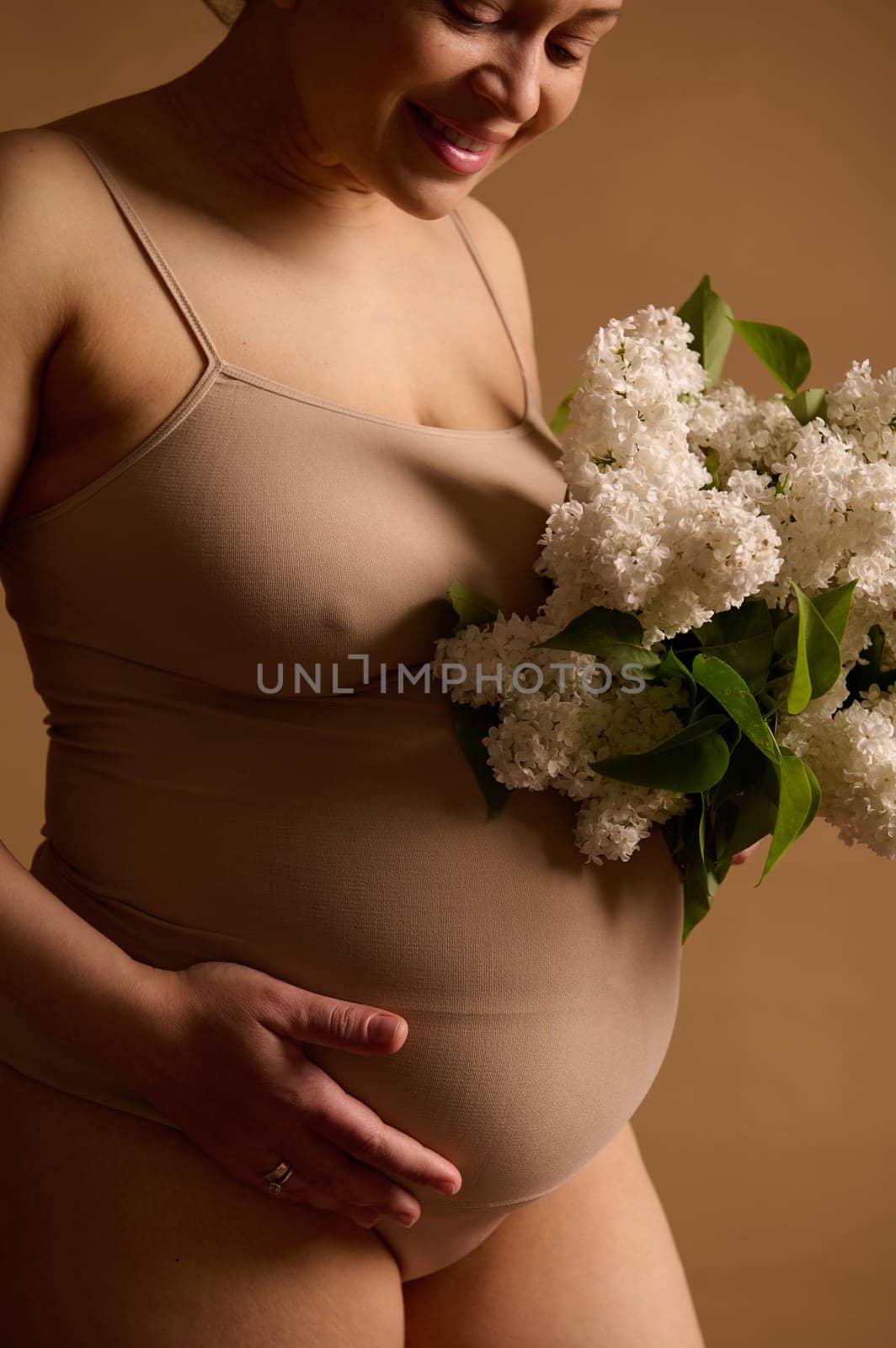 Close-up view smiling happy pregnant woman, expectant young mother in beige lingerie with bunch of white lilac flowers, touches and strokes her big belly in late pregnancy, isolated studio background