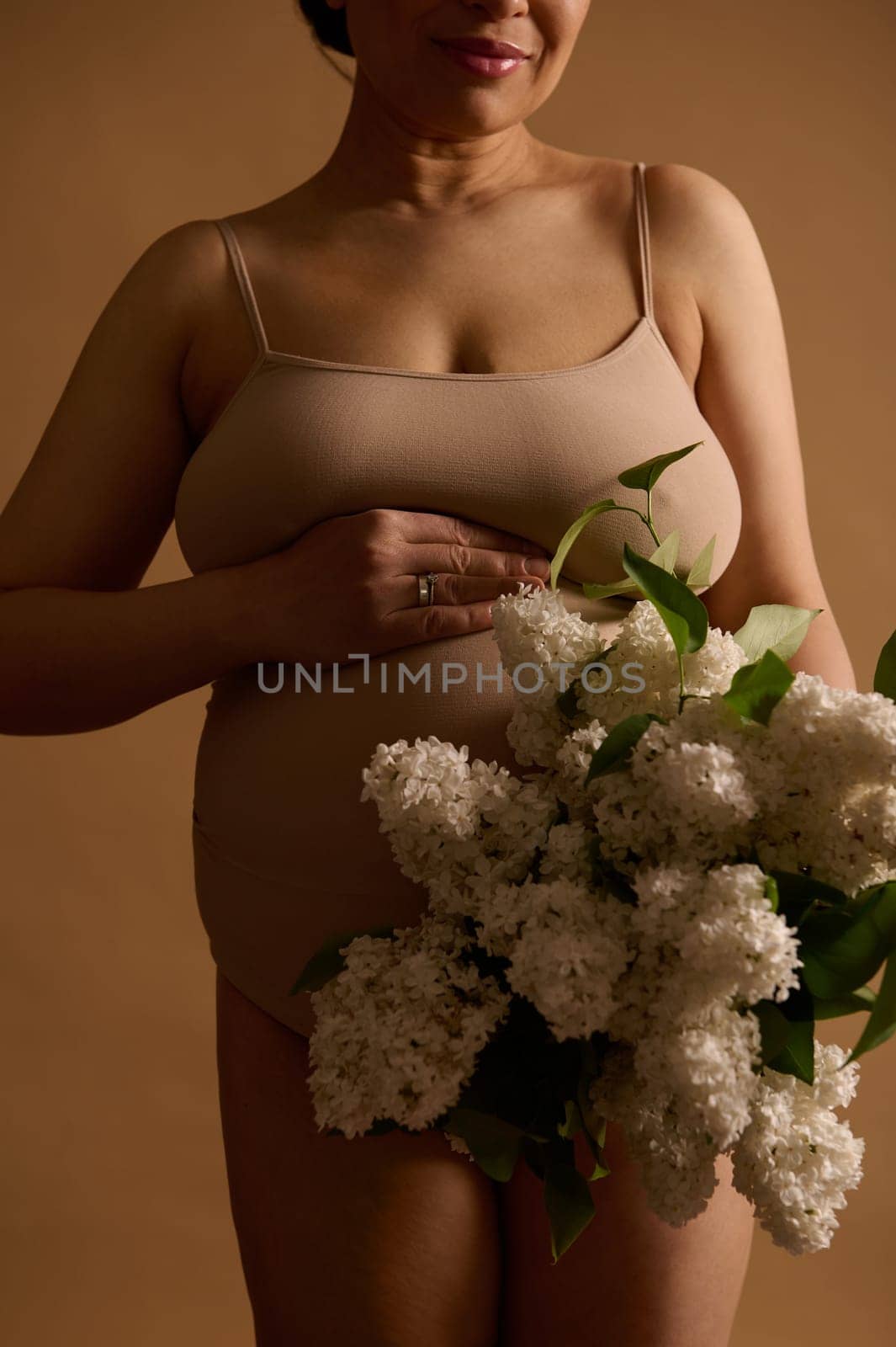 Midsection of pregnant woman holding a bouquet of white lilac, posing over beige background with her hands on belly. The concept of healthy happy pregnancy and maternity. Women's health and fertility