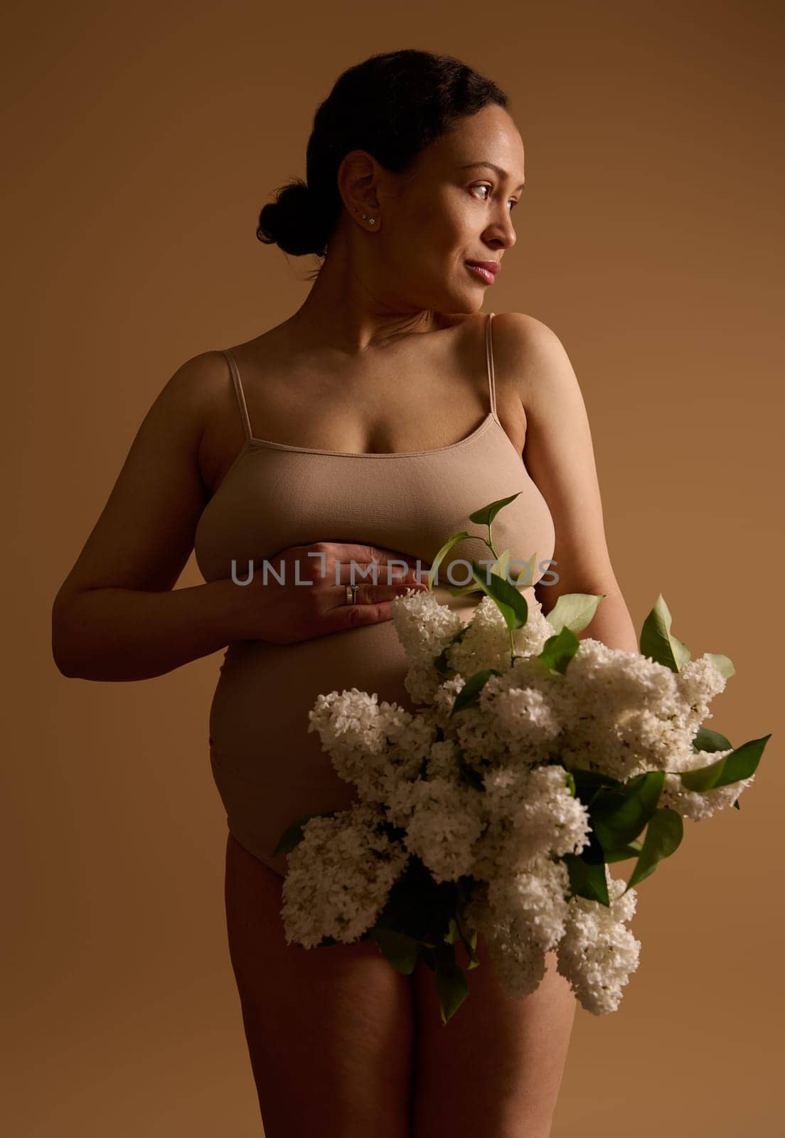 Gravid woman in beige underwear, holding hand on her pregnant belly, looking aside, posing with bunch of blooming lilacs by artgf