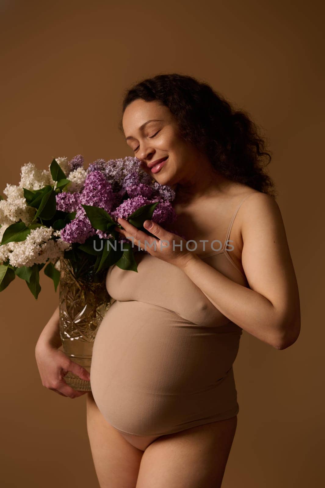 Delightful gravid pregnant woman in beige lingerie, posing with purple and while blooming lilacs, on beige background by artgf