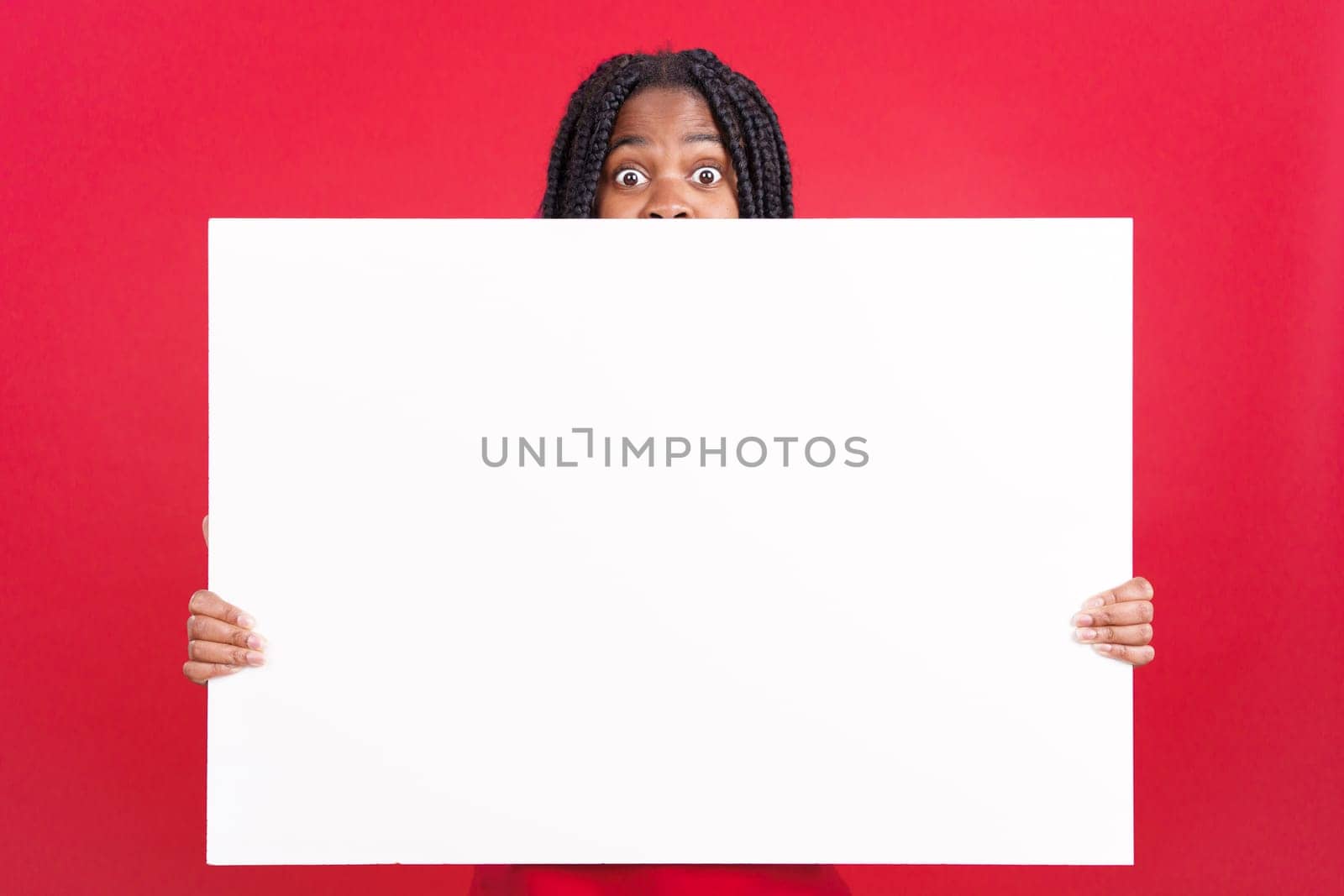 Surprised african woman hiding behind a blank panel in studio with red background