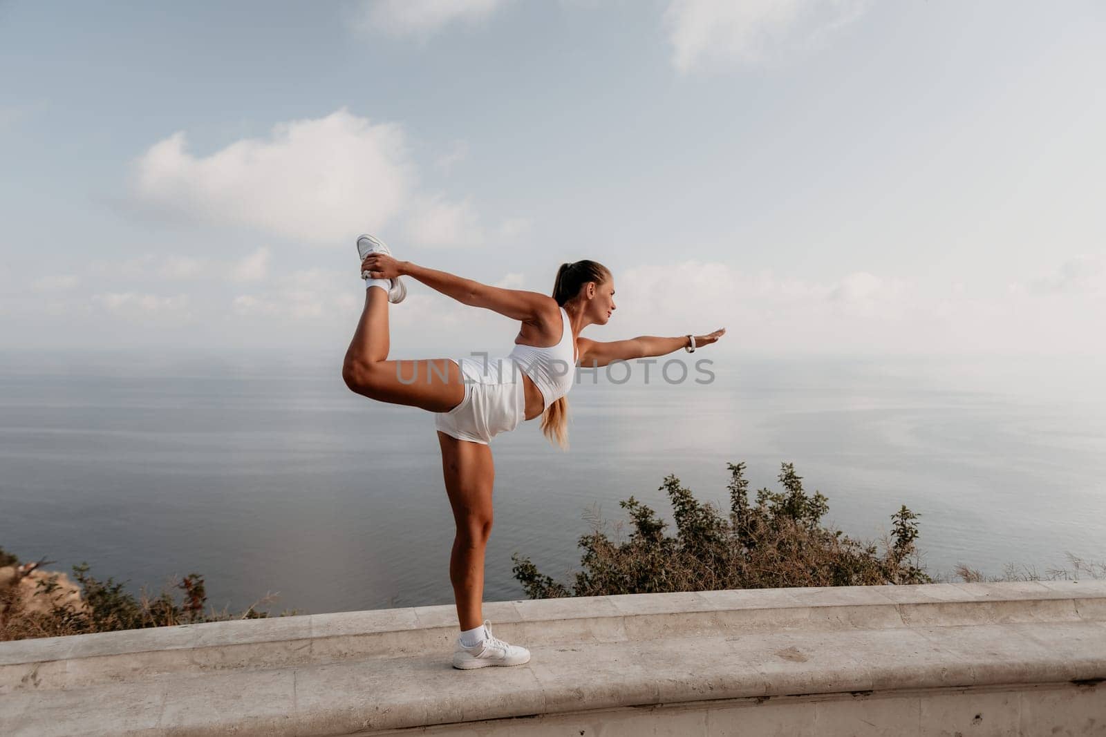 Fitness woman sea. A happy middle aged woman in white sportswear exercises morning outdoors in a park with a beach view. Female fitness pilates yoga routine concept. Healthy lifestyle. by panophotograph