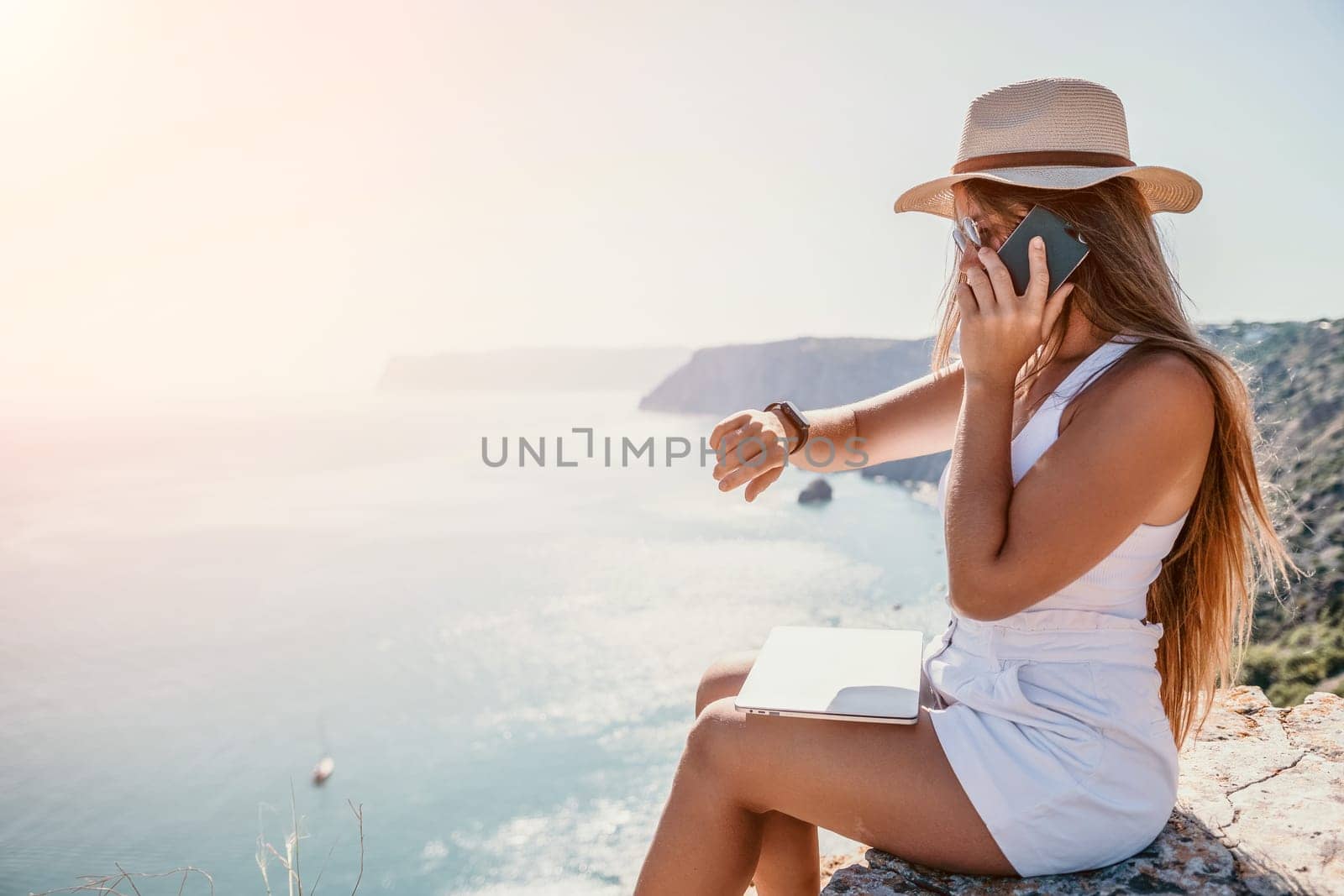 Successful business woman in yellow hat working on laptop by the sea. Pretty lady typing on computer at summer day outdoors. Freelance, travel and holidays concept.