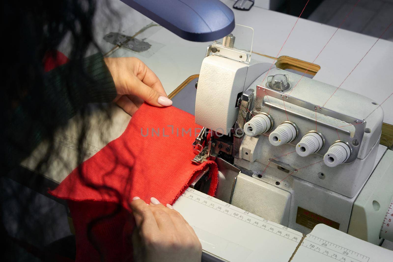 Young woman using professional overlock sewing machine in workshop. Equipment for edging, hemming or seaming clothes at tailors shop