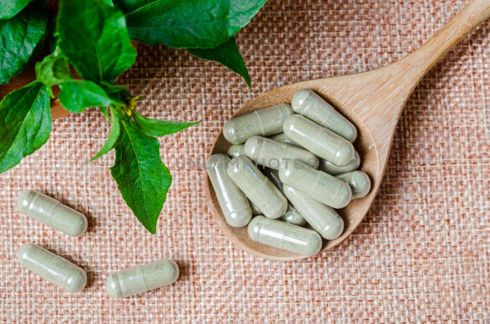 Pile of herbal medicine capsules in wooden spoon with green leaf on sack background.