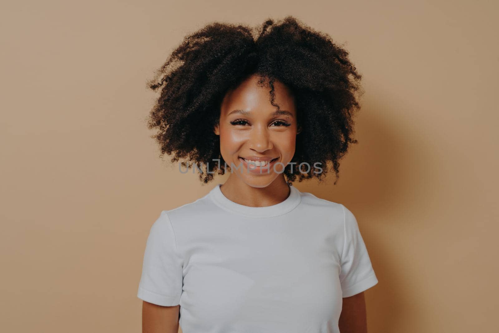 Portrait of happy young african woman with broad shining smile dressed in white tshirt posing alone against beige background, positive mixed race female being in high spirit. Positive people concept