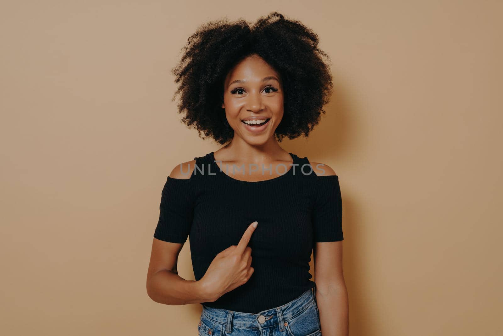 Young mean me? Portrait of happy young mixed race woman pointing finger at herself and looking at camera with amazed face expression, dressed in black t shirt. Body language and human emotions