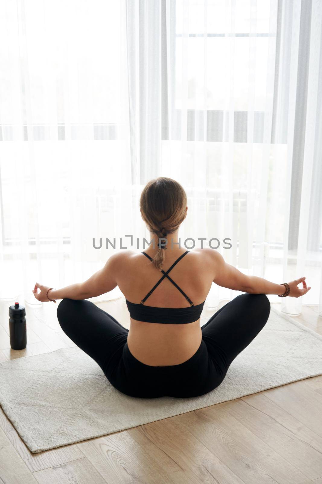 Rear view of girl relaxing in lotus pose at home in front of window.Yoga concept by Mariakray