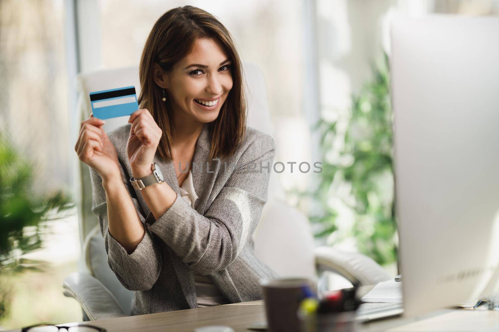 An attractive business woman holding a credit card and looking at camera while working on her computer in the office.