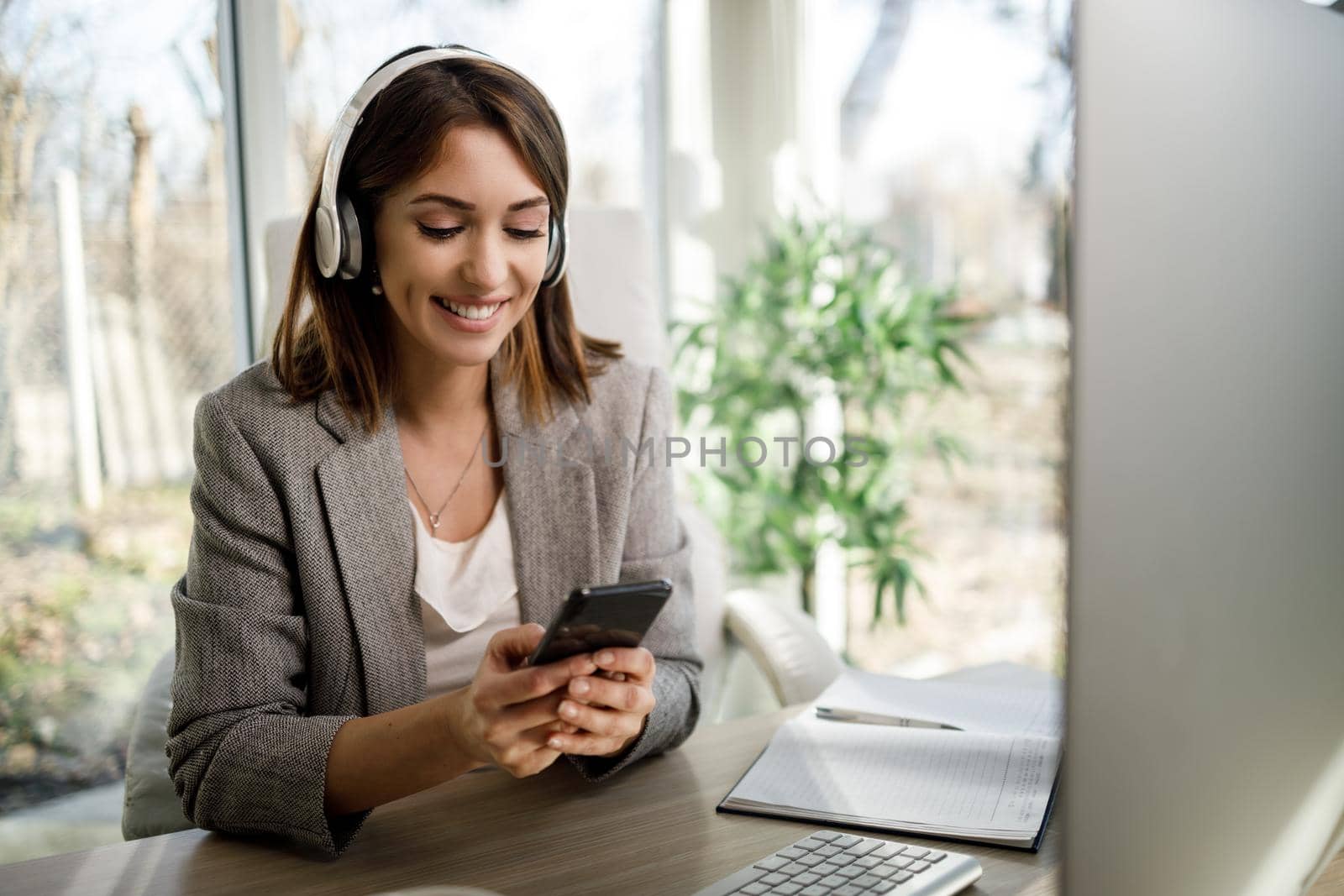 A smiling young woman with headphones using smartphone in her home office.