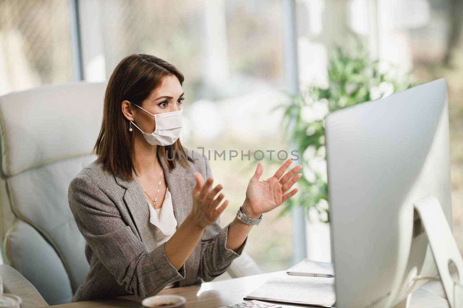 An attractive young woman with surgical mask sitting alone in her office and making video call on computer. 