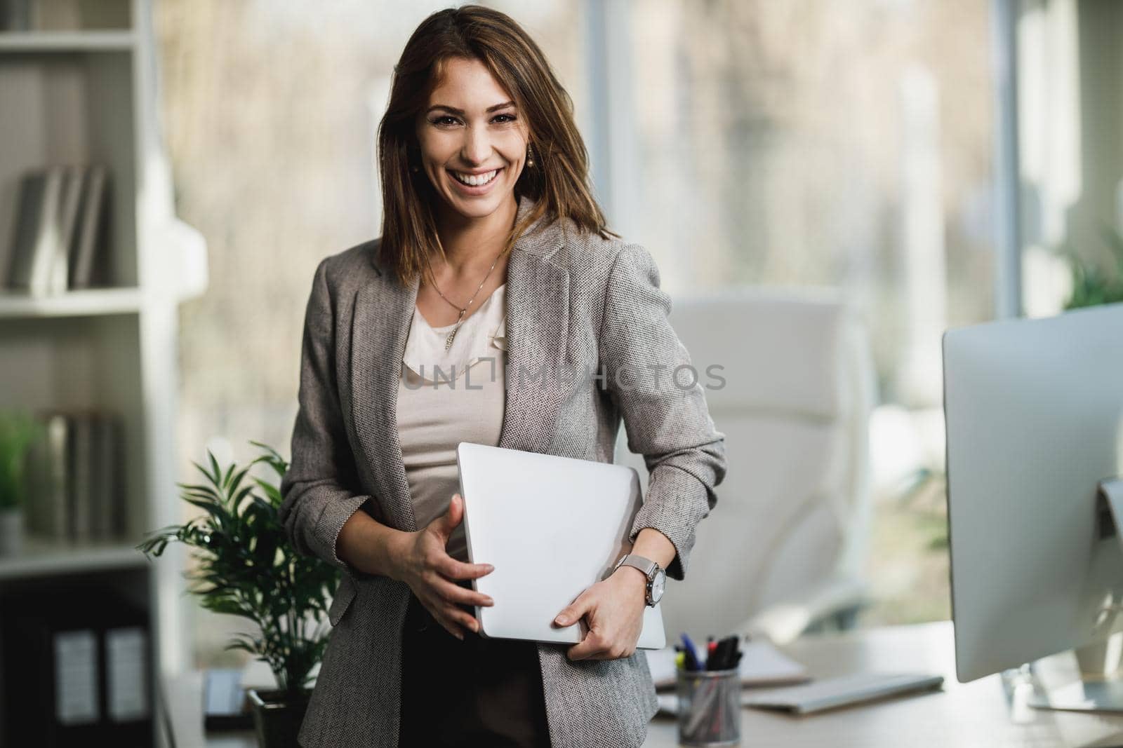 A successful young business woman holding laptop and standing in her office. Looking at camera.