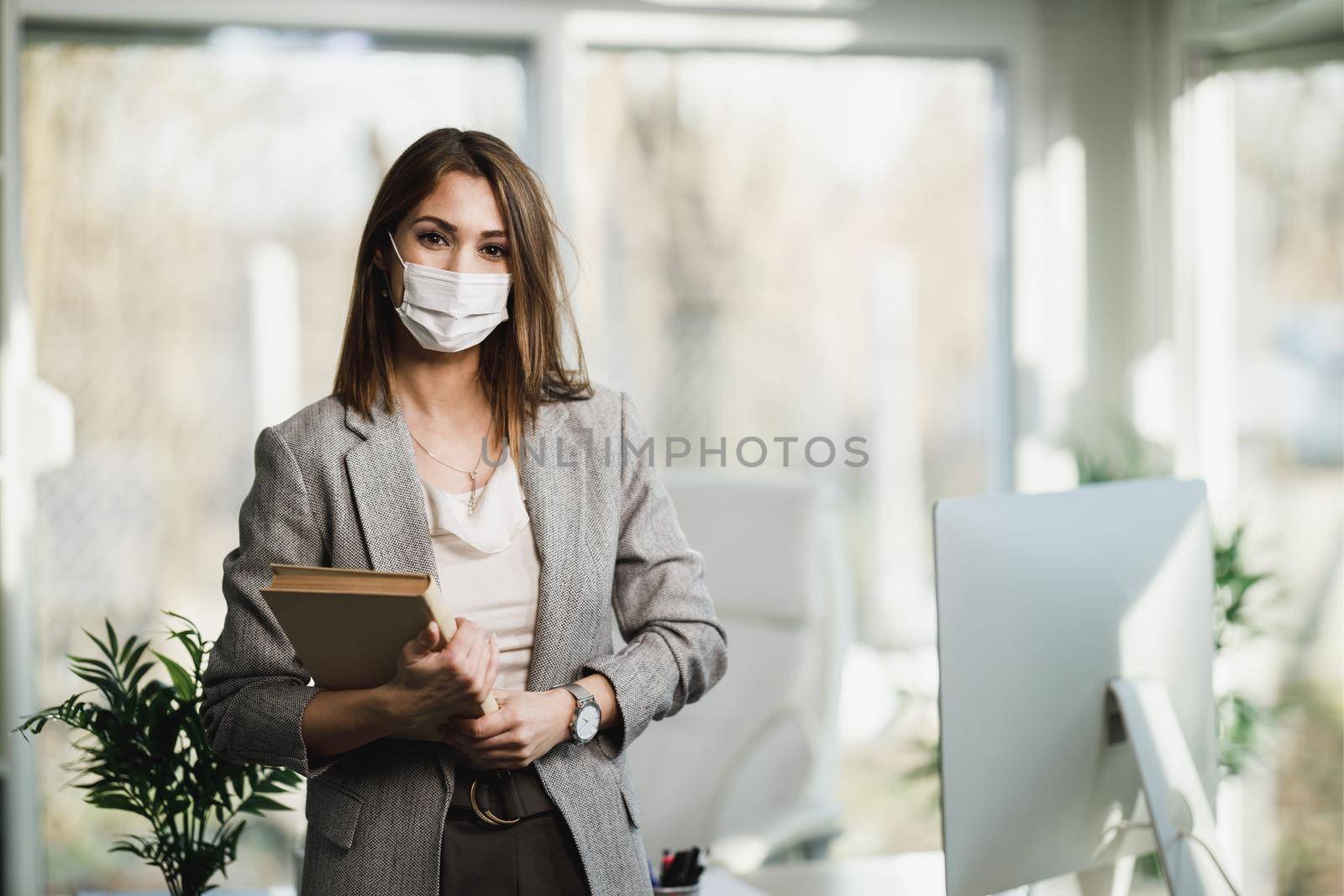 A successful young business woman with surgical mask standing in her office. Looking at camera.