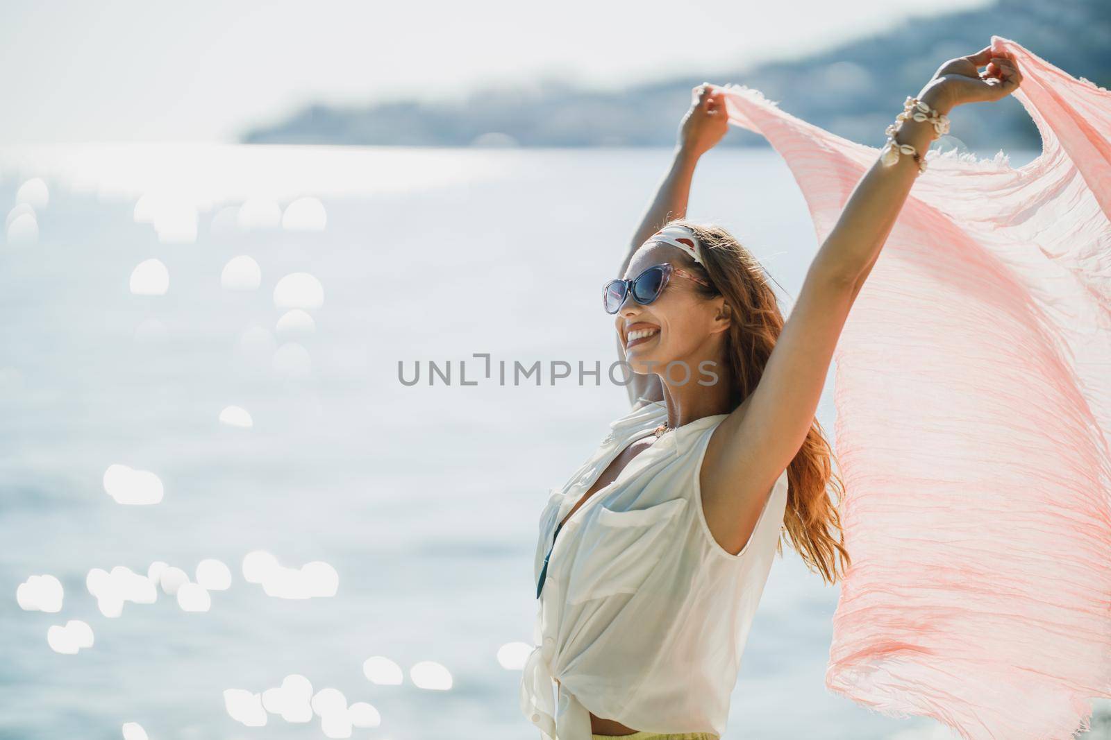 An attractive young woman is having fun and enjoying a summer vacation. She is posing with transparent scarf in her hands on the beach.