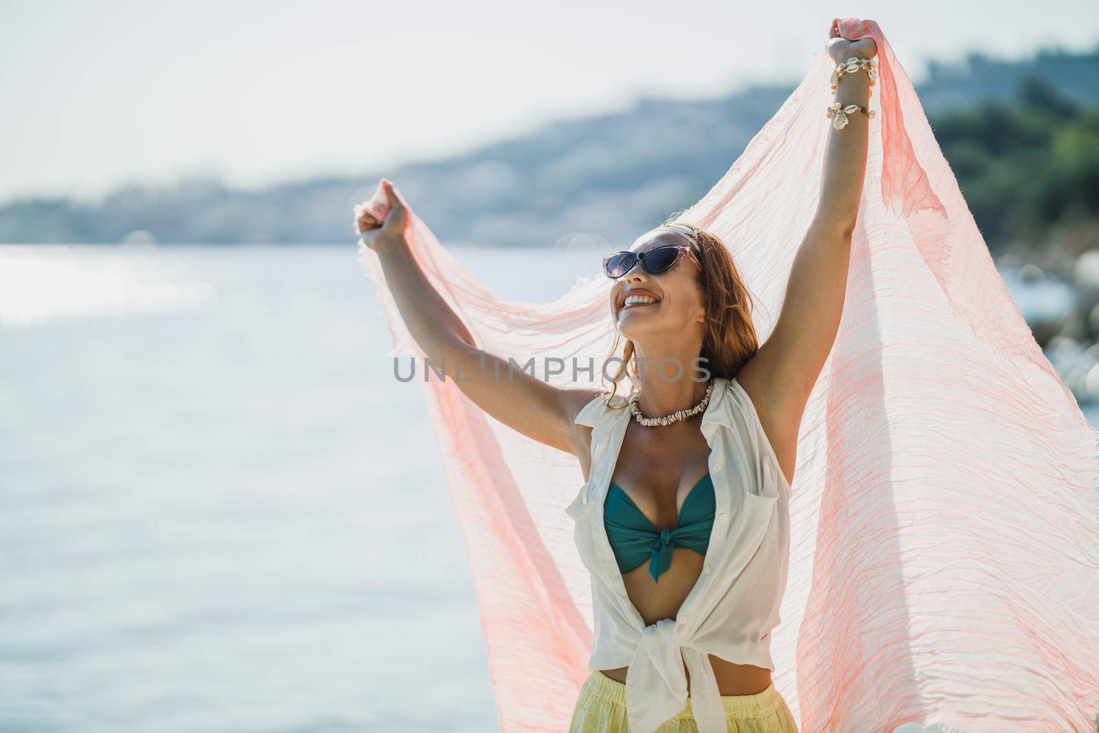 An attractive young woman is having fun and enjoying a summer vacation. She is posing with transparent scarf in her hands on the beach.