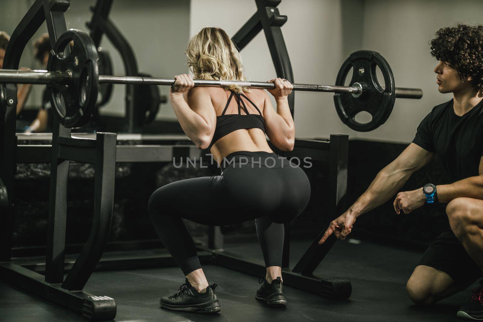 A young muscular woman is doing exercises for her legs supporting by coach in the gym.