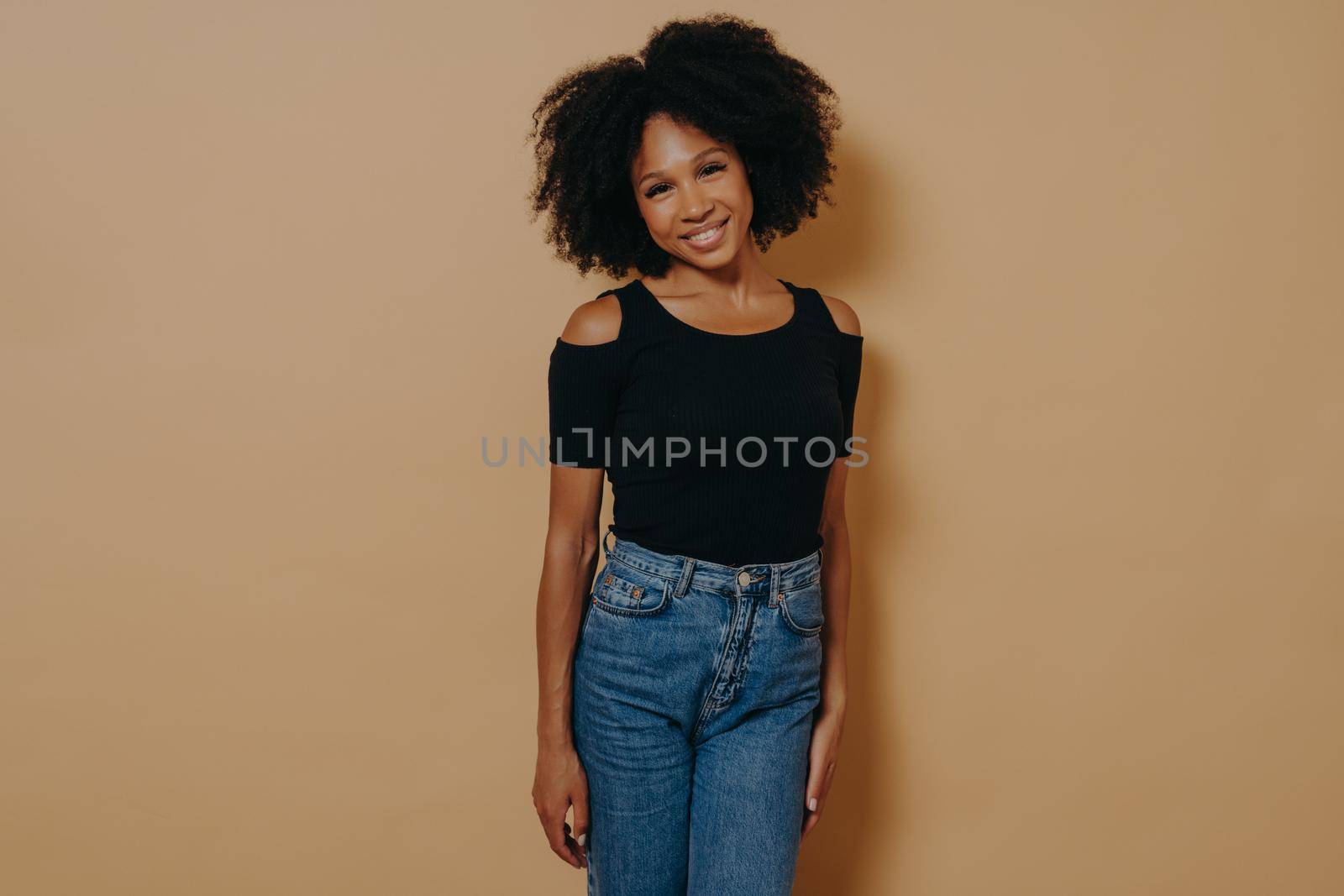 Indoor photo of pretty afro american smiling woman in casual outfit looking at camera with shy charming smile, posing isolated over dark beige background. Beautiful dark skinned women