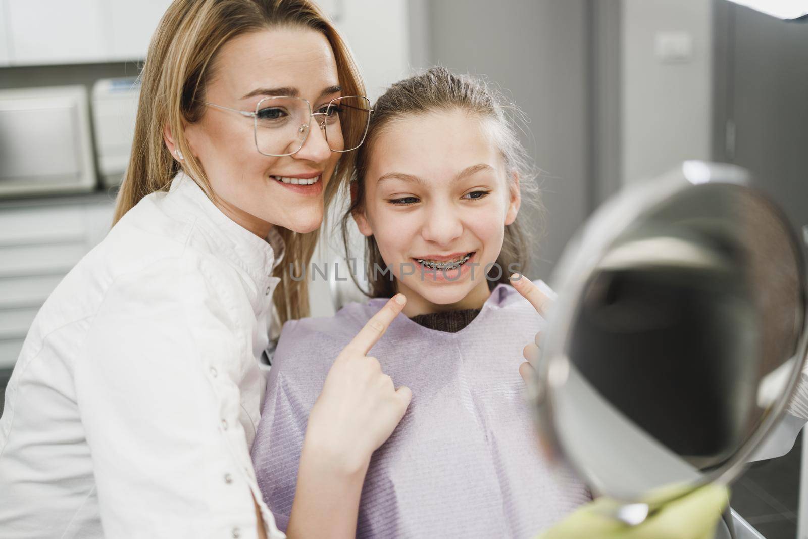 A happy teenager girl looking her teeth in a mirror after dental procedure at dentist's office.