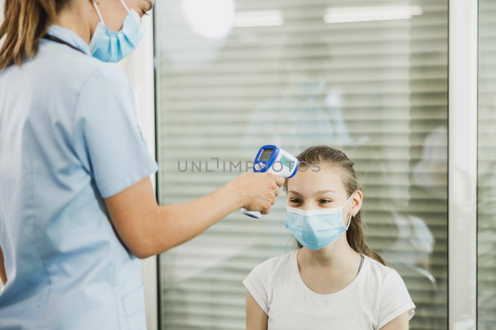 Nurse measuring temperature of a cute female teenager in waiting room during coronavirus pandemic.
