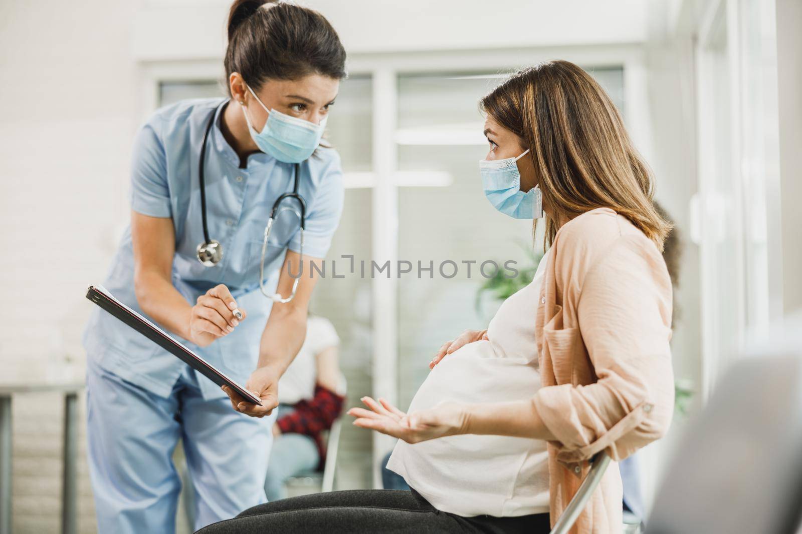 Nurse talking to worried pregnant woman and analyzing medical report before covid-19 vaccine at hospital waiting room.