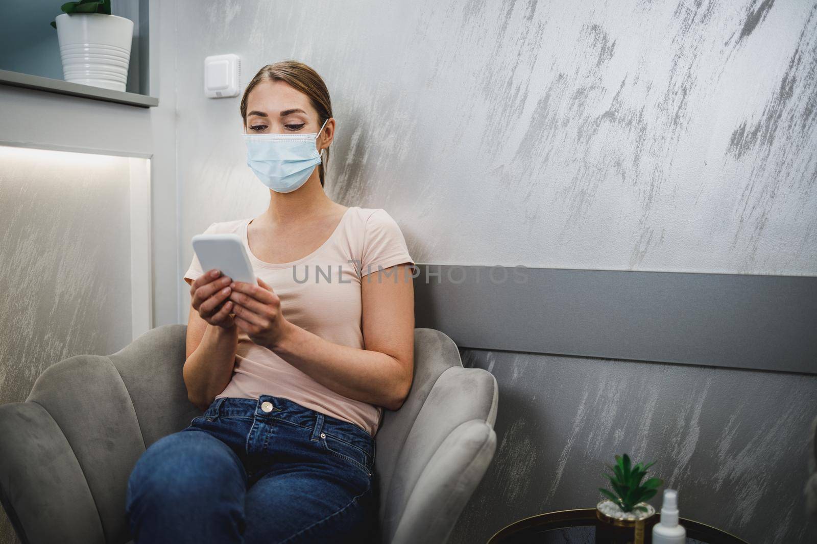 An attractive young woman wearing face mask and using her cellphone while sitting on a chair in waiting room at dentist's office.