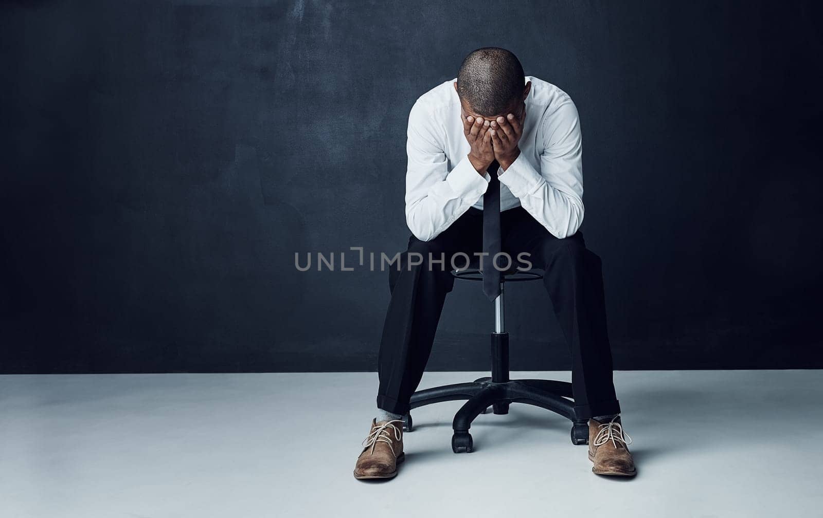 Everyone has battles to face before they see success. Studio shot of a young businessman sitting with his hands covering his face against a dark background. by YuriArcurs