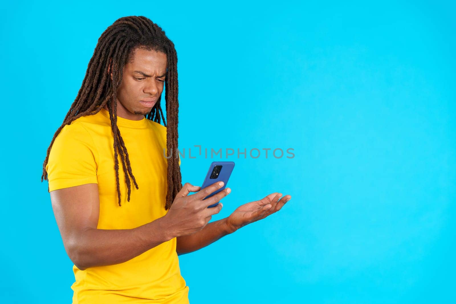 Worried latin man with dreadlocks using a mobile phone in studio with blue background