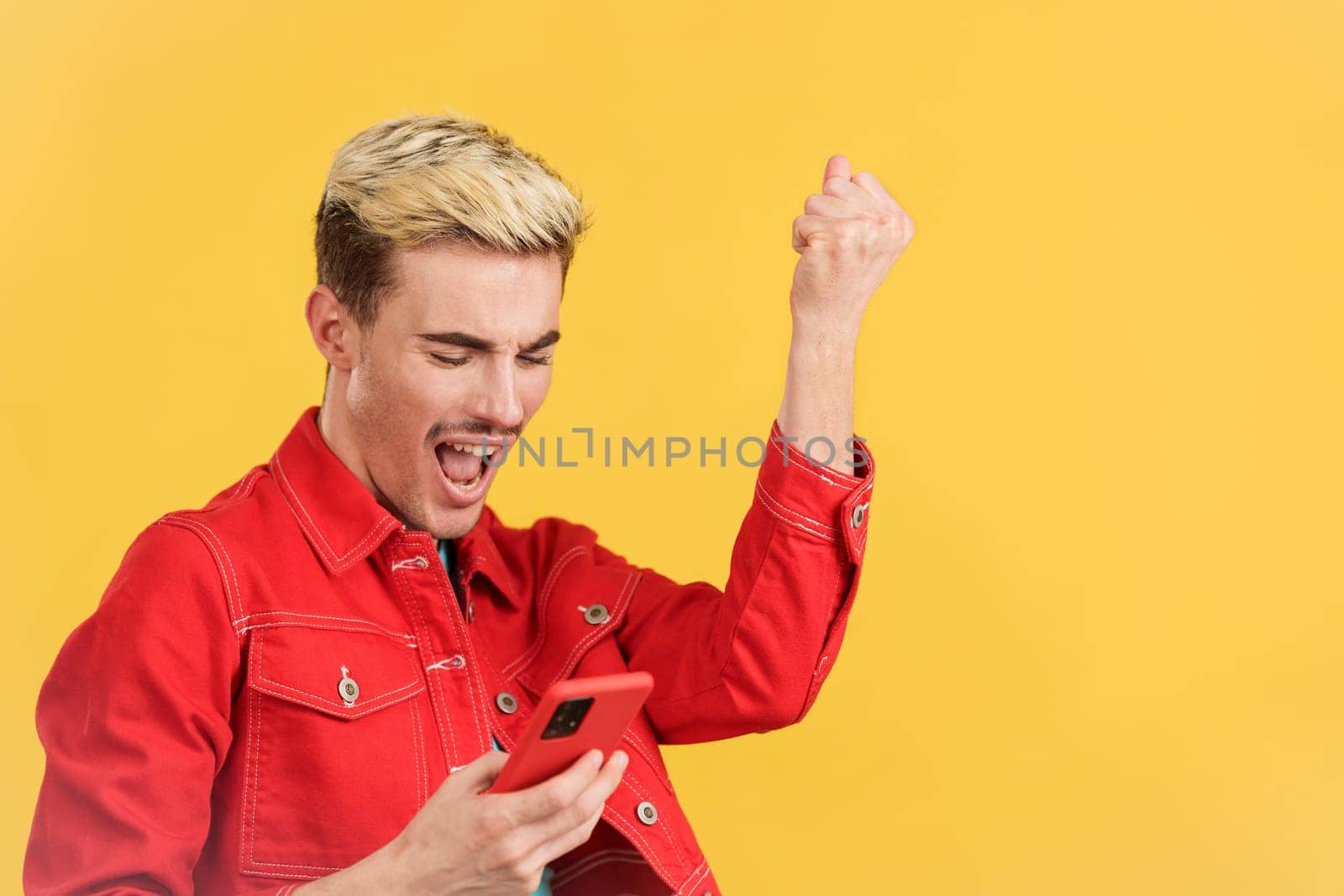 Excited gay man celebrating while using the mobile phone in studio with yellow background