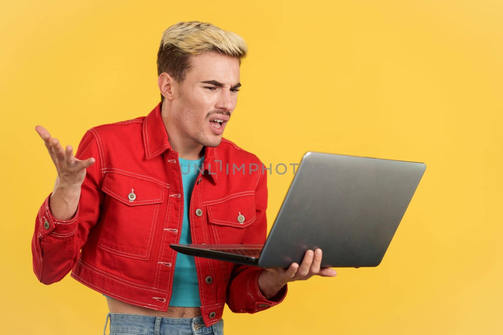 Gay man gesturing while having a problem with a laptop in studio with yellow background