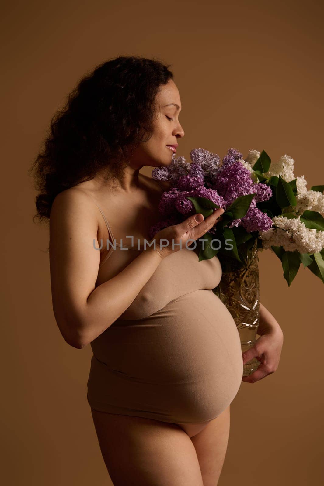 Attractive gravid woman, pregnant mother in beige lingerie, holding bouquet of blooming lilacs, isolated background. by artgf
