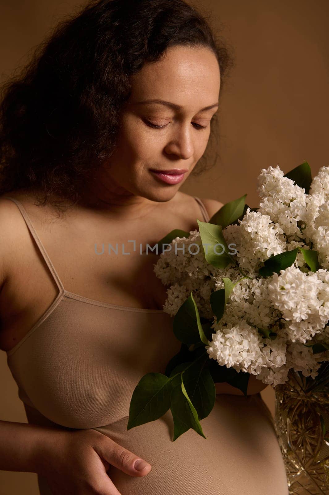 Close-up studio portrait of beautiful multiethnic gravid woman, pregnant expectant mother in beige lingerie, holding bouquet of blooming lilacs, isolated background. Pregnancy fashion. Body positivity