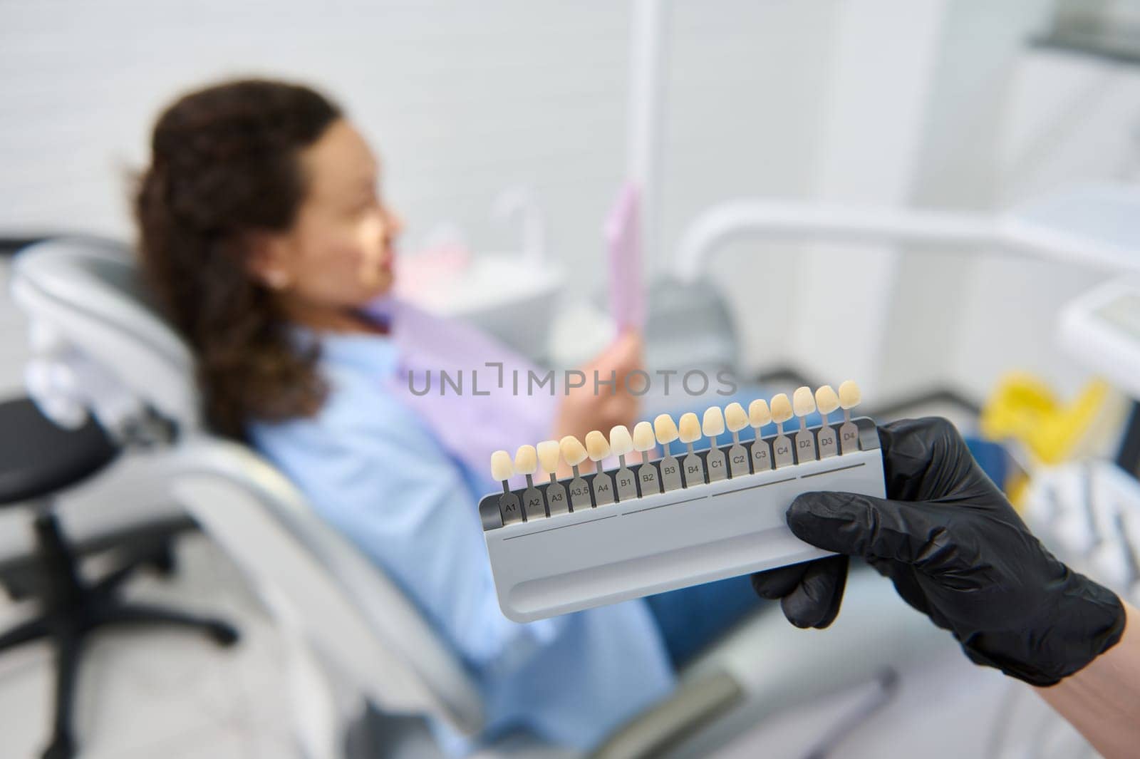 Vita scale. Bleaching teeth guide. Tooth color chart in the hands of a dentist orthodontist doctor, against blurred background of a female patient sitting in dental chair in modern dentistry clinic