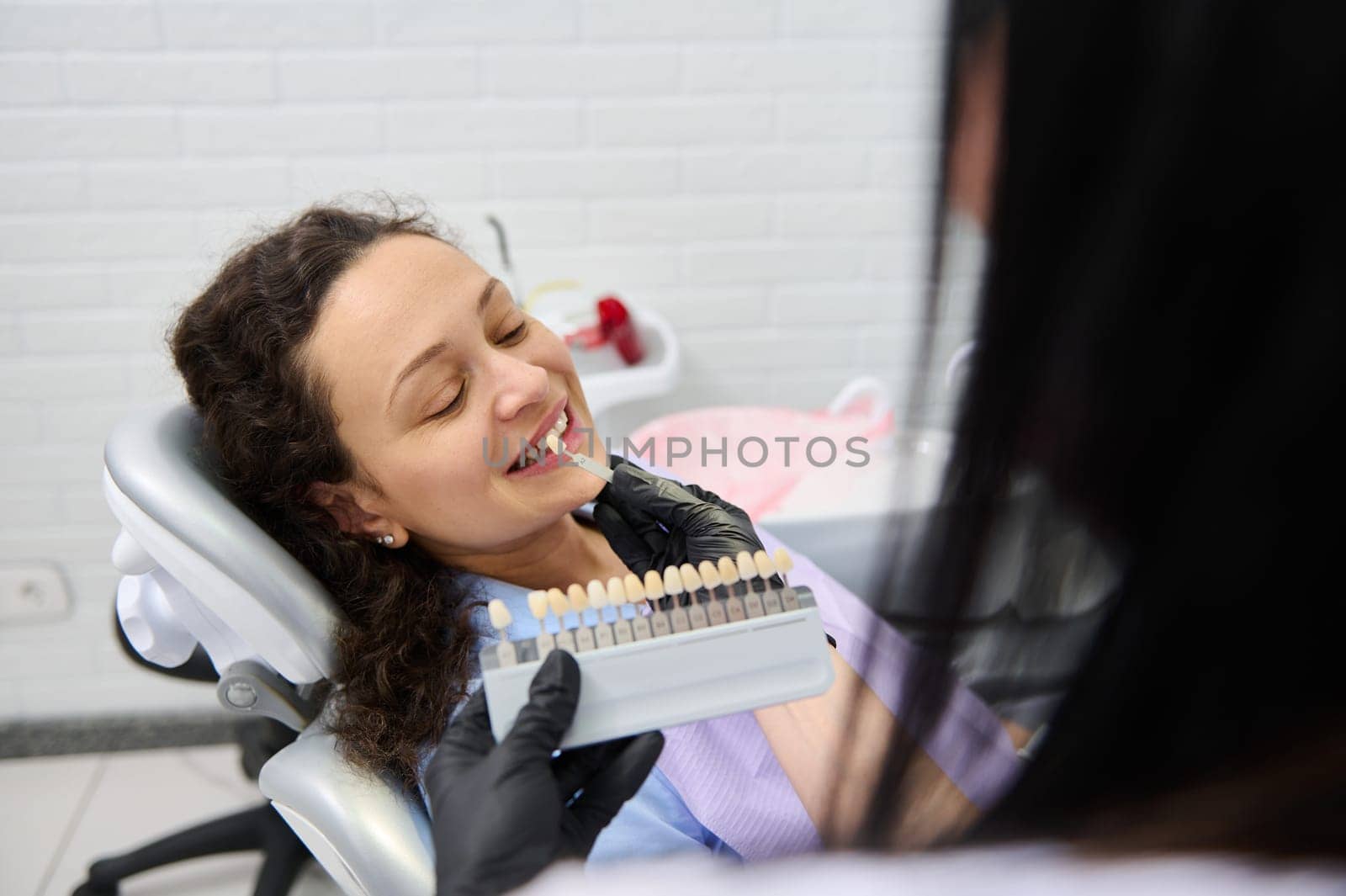 Dentist doctor placing tooth color chart, over a beautiful smiling woman's teeth, choosing the shade of veneers, according to Vita scale. Visual method of tooth color subjective perception. Dentistry