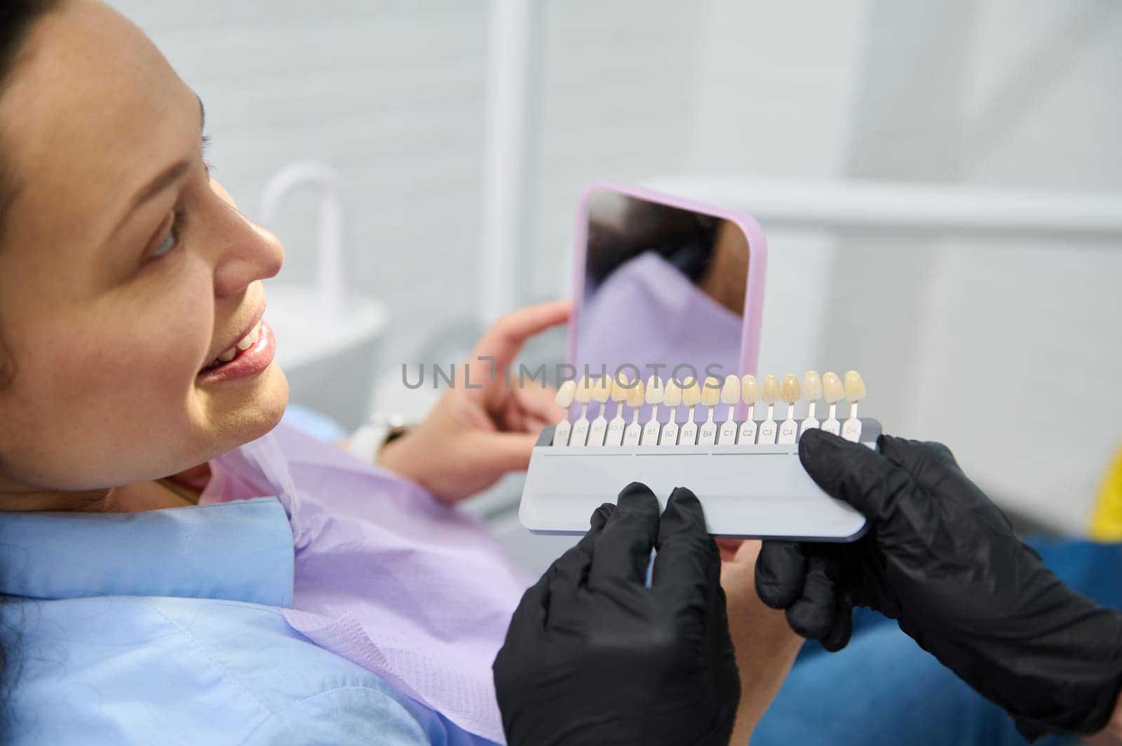 Smiling female patient receives dental care, teeth bleaching. Dentist checks tooth color using verneer Vita scale chart by artgf