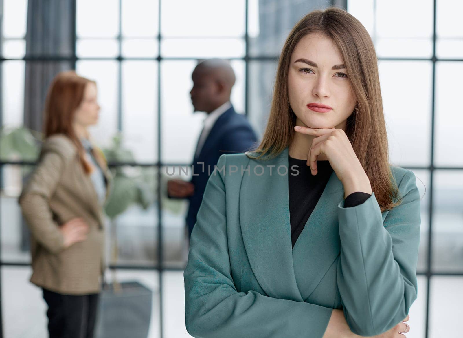 businesswoman standing with her arms crossed in an office while her colleagues have a meeting in the background.