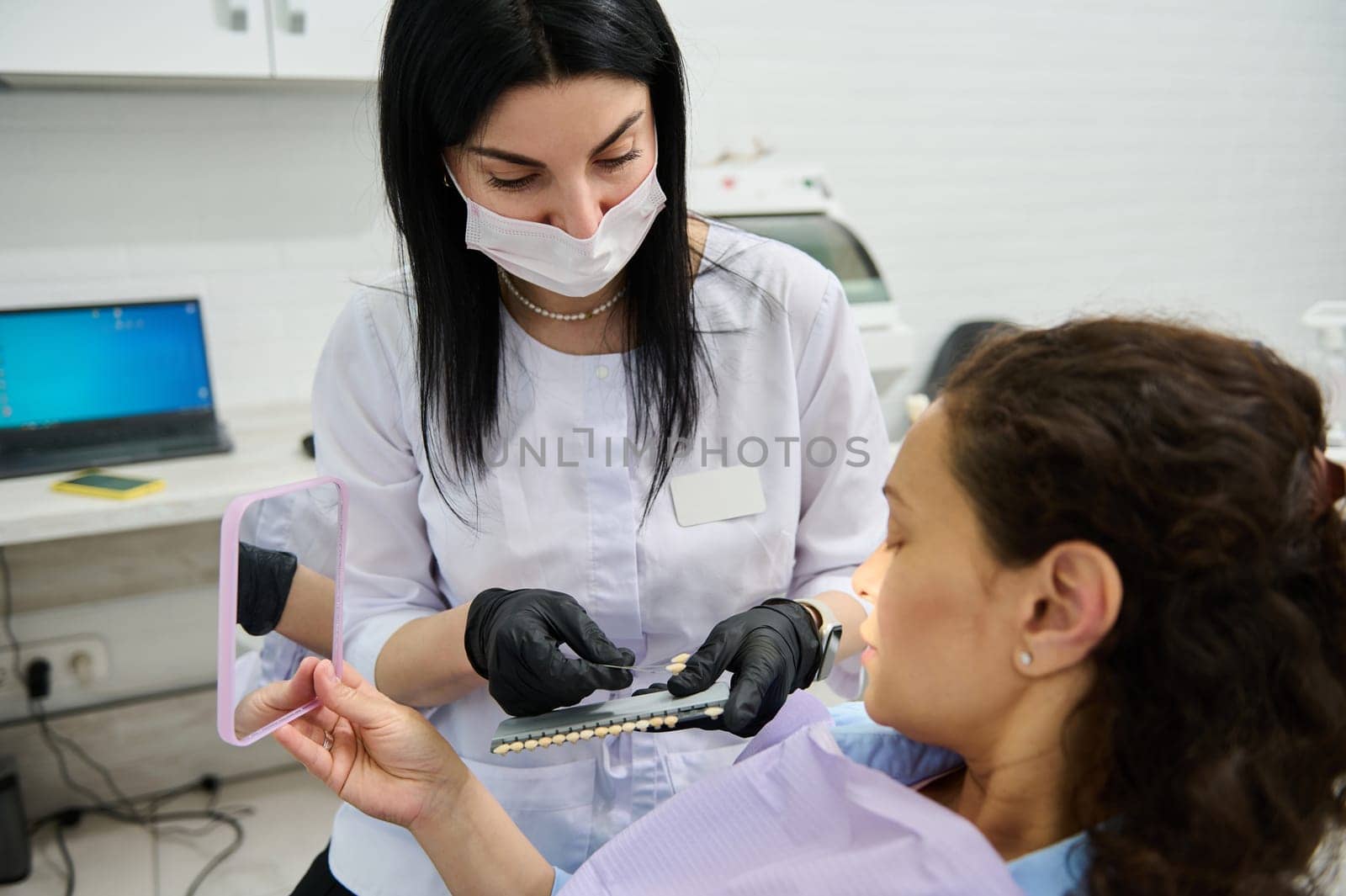 Female dentist doctor and her patient during medical checkup in dentistry clinic. Woman sitting in dentist's chair and smiling looking at mirror while visiting dentist for teeth bleaching procedure