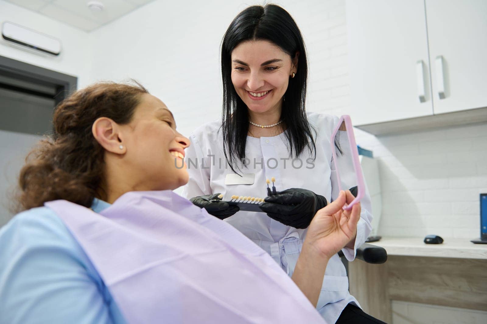 Dental surgeon comparing female patient teeth with shade guide according to Vita scale. Smiling happy woman looking at her mirror reflection after teeth whitening procedure in dentistry clinic