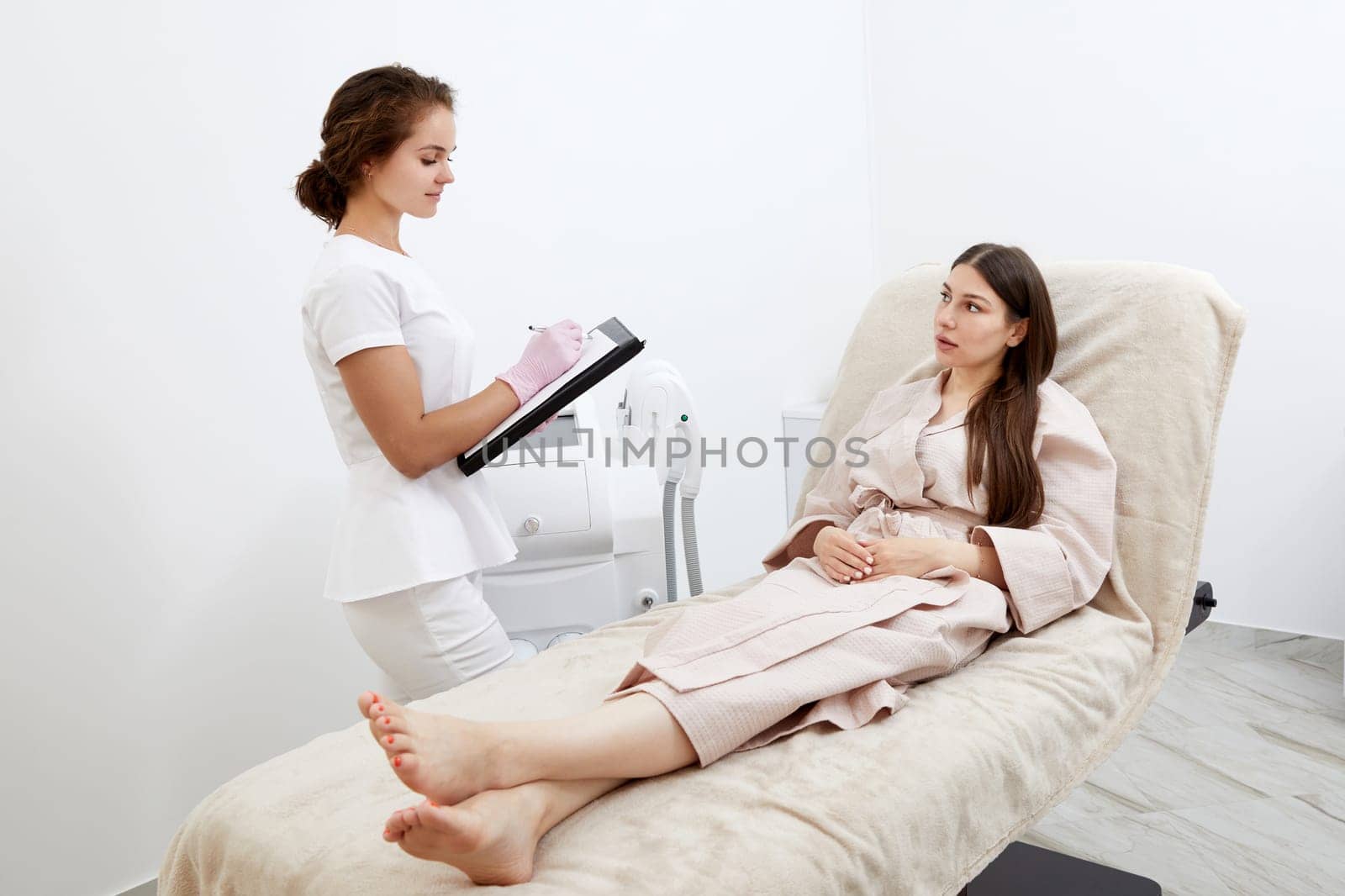 Professional cosmetician woman with papers talking to woman before procedure in clinic of esthetic cosmetology by Mariakray