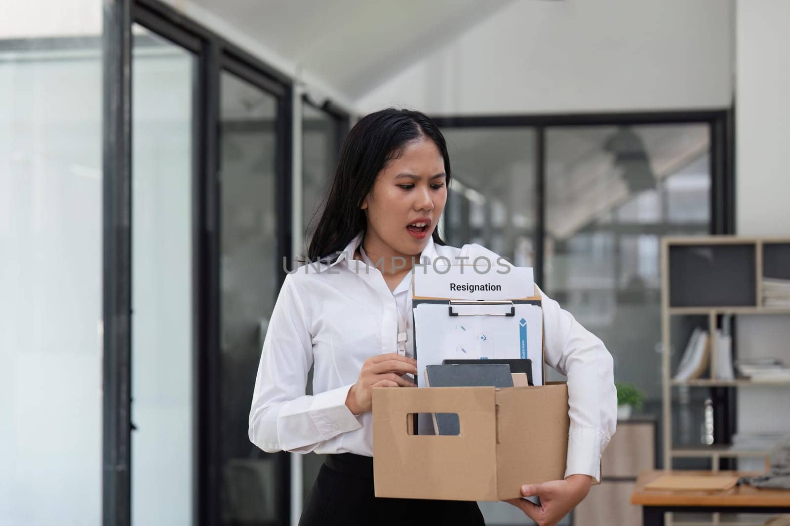 An excited young Asian female employee feels happy to quit her job, holding a cardboard box with her stuff and resignation letter while standing at office.